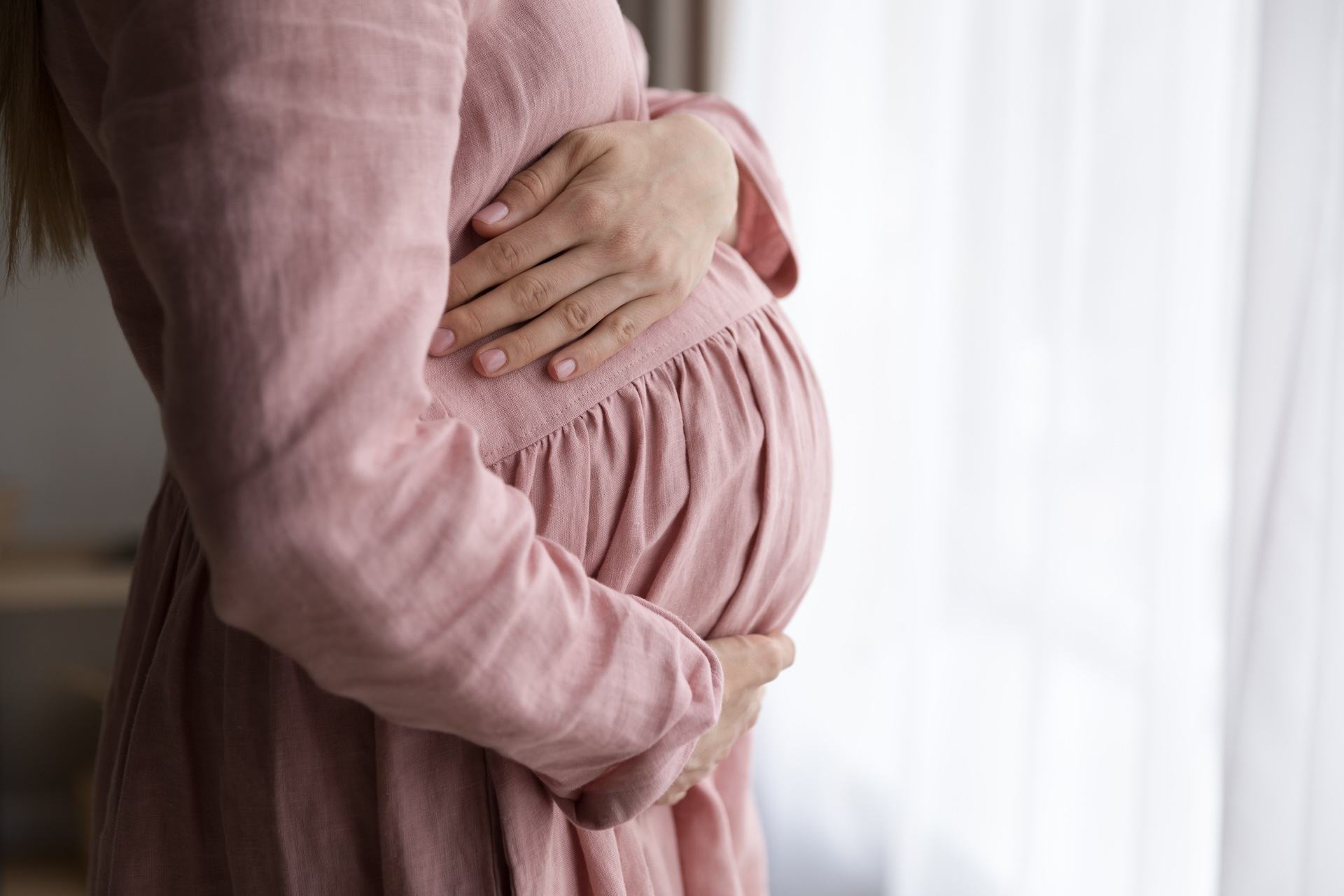 Uma mulher grávida em um vestido rosa segura a barriga enquanto olha pela janela