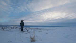 Um homem com roupas de inverno está em um campo ártico com alguns pedaços de grama aparecendo na neve